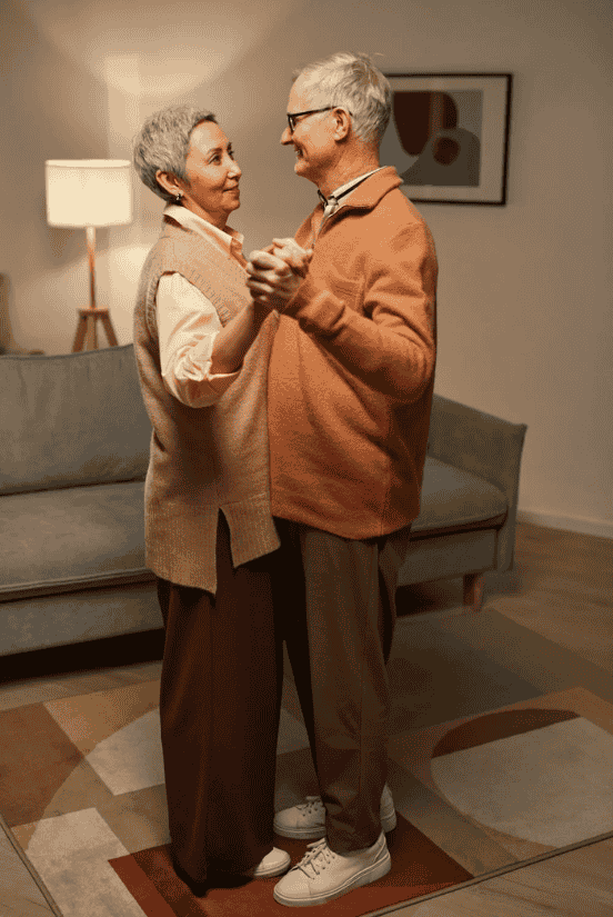 An elderly couple dressed in warm tones dances together in a cozy living room.